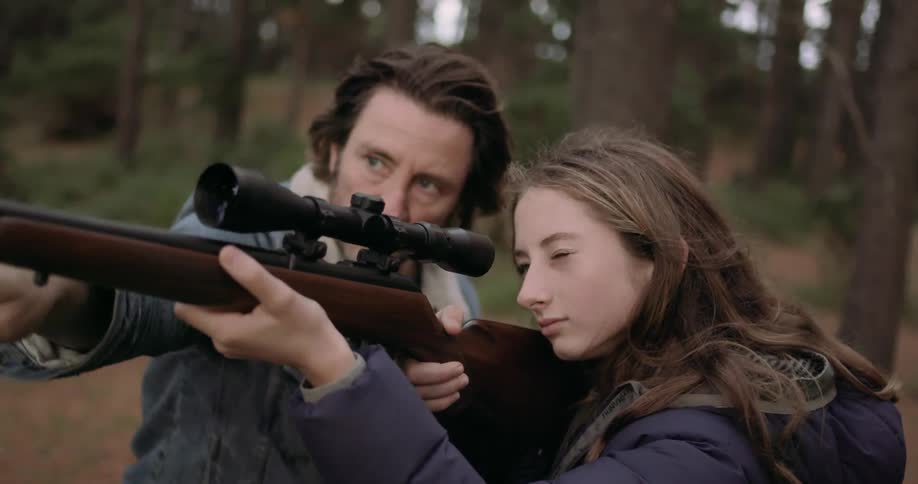 A young girl is holding a rifle and a man is showing her how to shoot