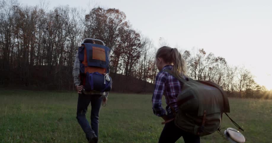 Man and girl wearing backpacks hike across a grassy field towards trees