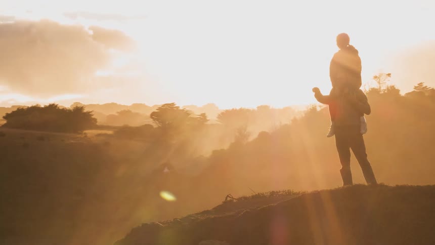 Child sitting on top man's shoulders walking outside on a hill at sunrise