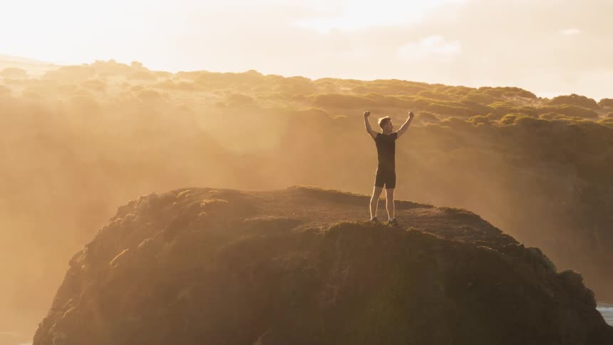 A man stands on a coastal cliff and raises his arms in the air