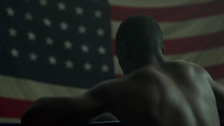 A shirtless man sits in front of a flag