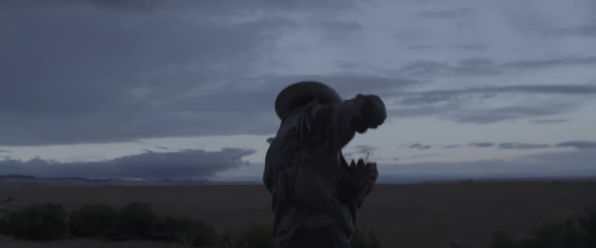 A cowboy walks across a grassy rural field at dusk