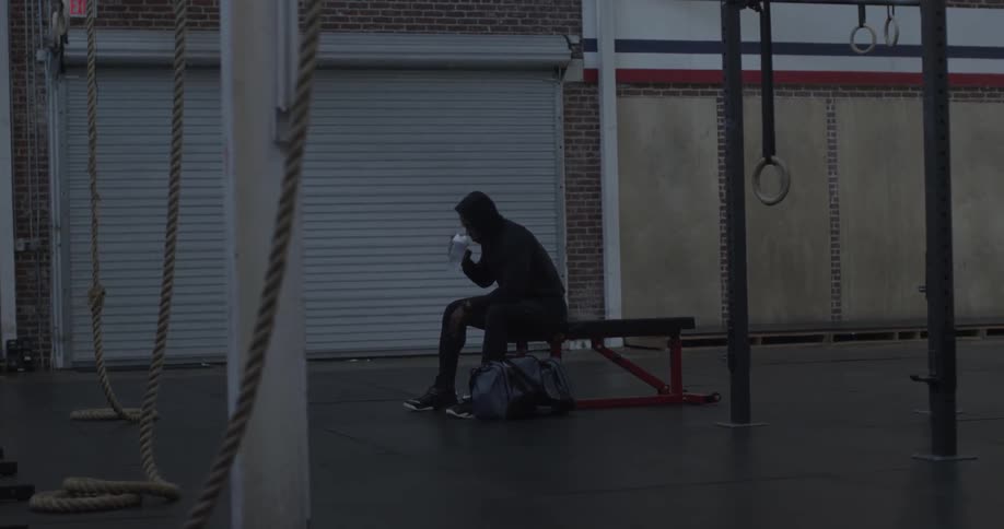 A man is sitting on the edge of a bunch in a hoodie drinking water