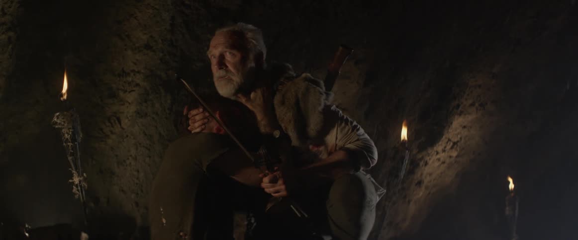 An old man comforting a bloodied man in a cave looks at something in the distance