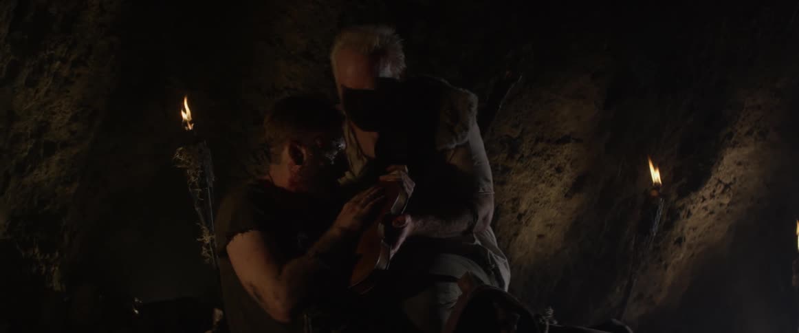 An elderly man tends to a hurt young man in a cave