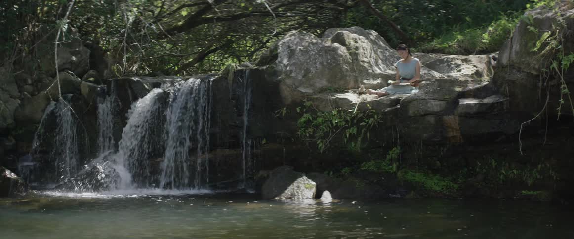 A woman sits on a rock ledge by a small river waterfall