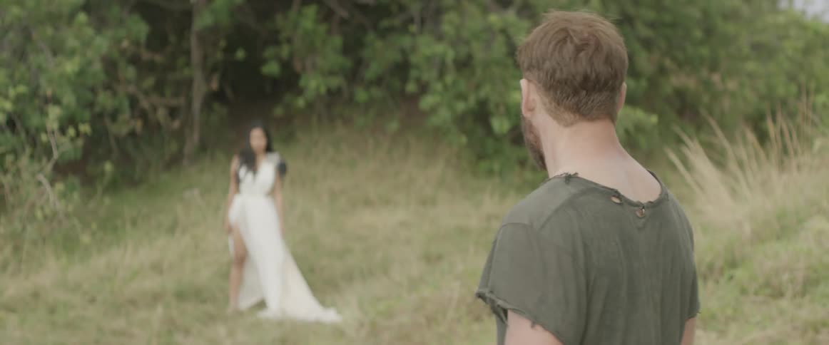 Woman in white gown walks away as a man walks towards her in a wooded area