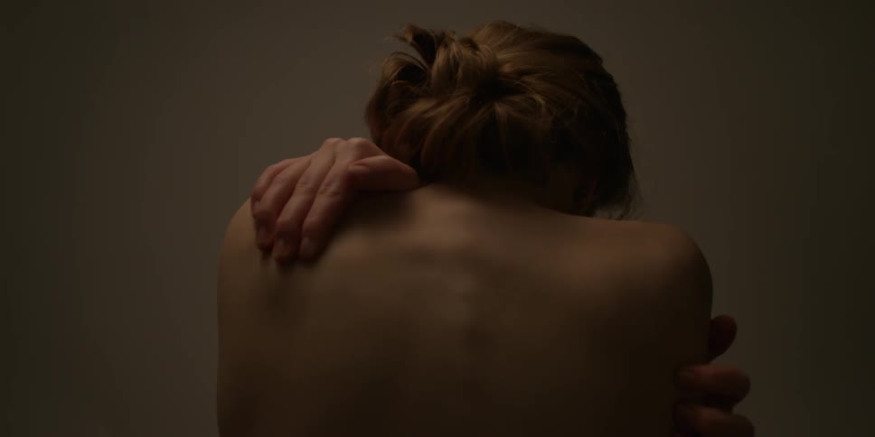 A shirtless woman wraps her arms around her shoulders and sways