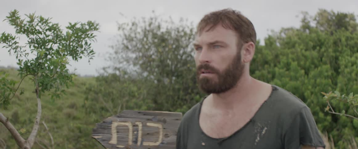 A bearded man follows a sign in a wooded area