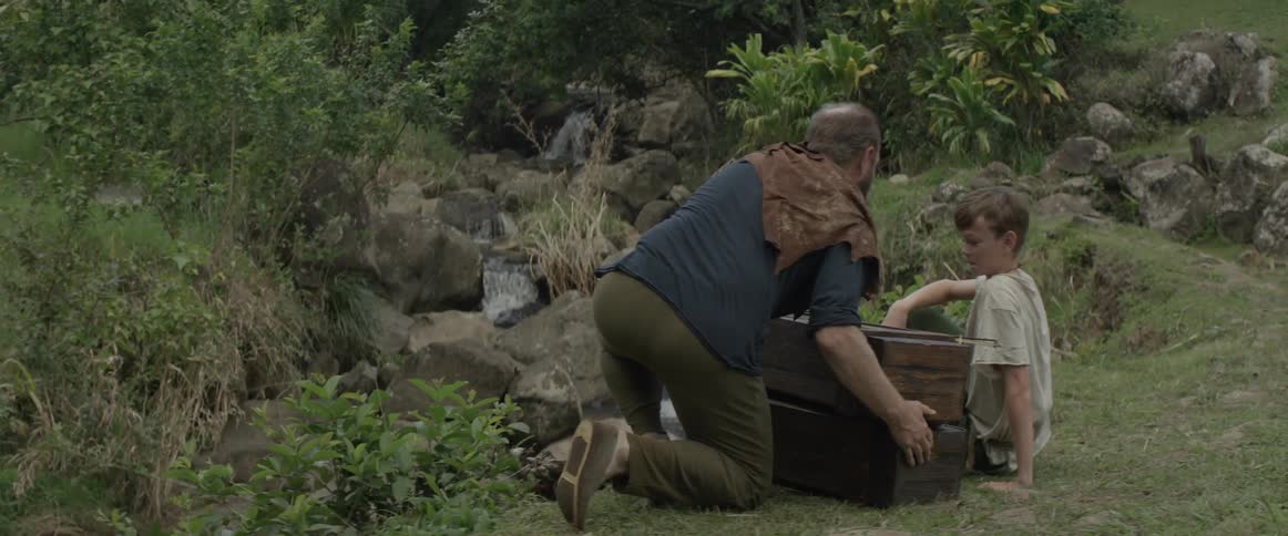A man places two wooden boxes next to his son sitting in the grass