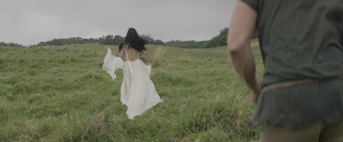 Woman in white dress smiling and running through grassy field as man follows her