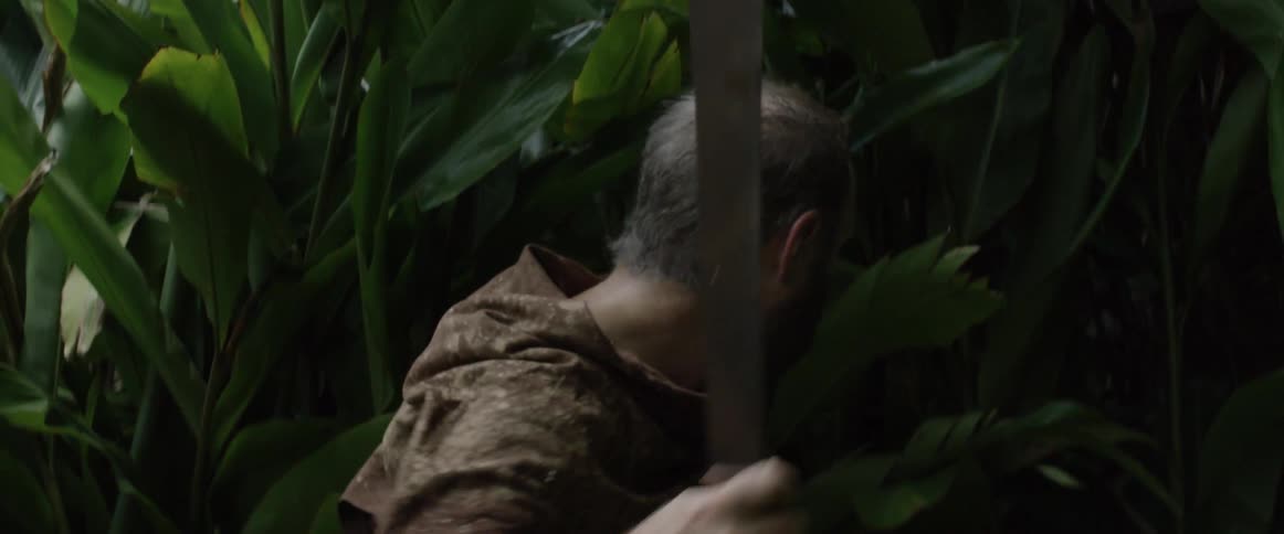 A man walks through thick brush swinging a machete