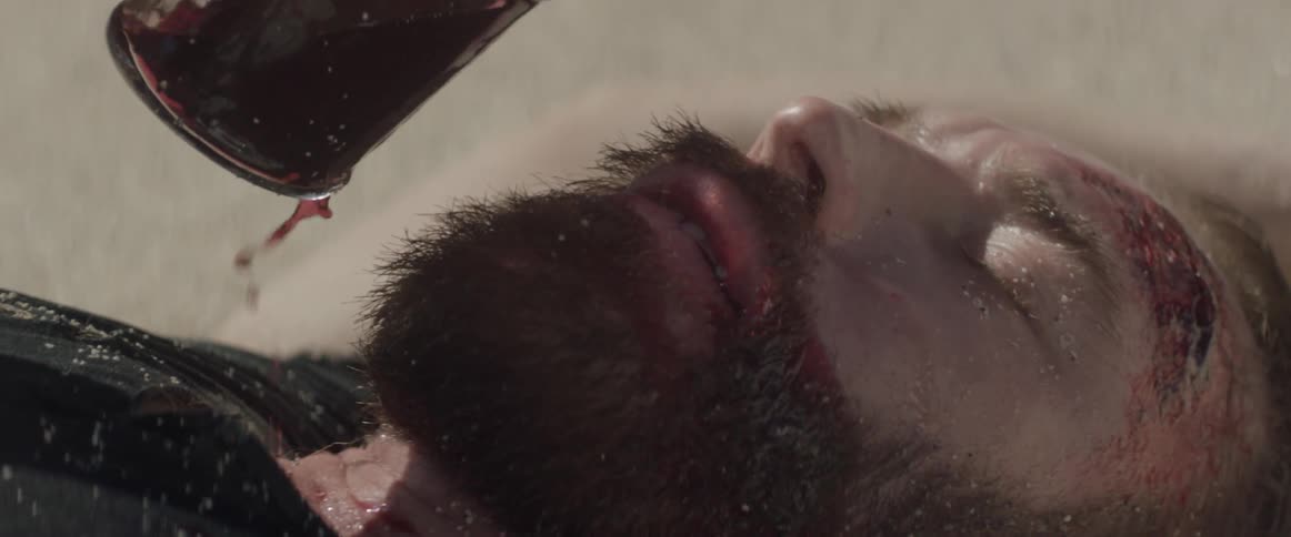 Sand covered man pours red liquid into his mouth