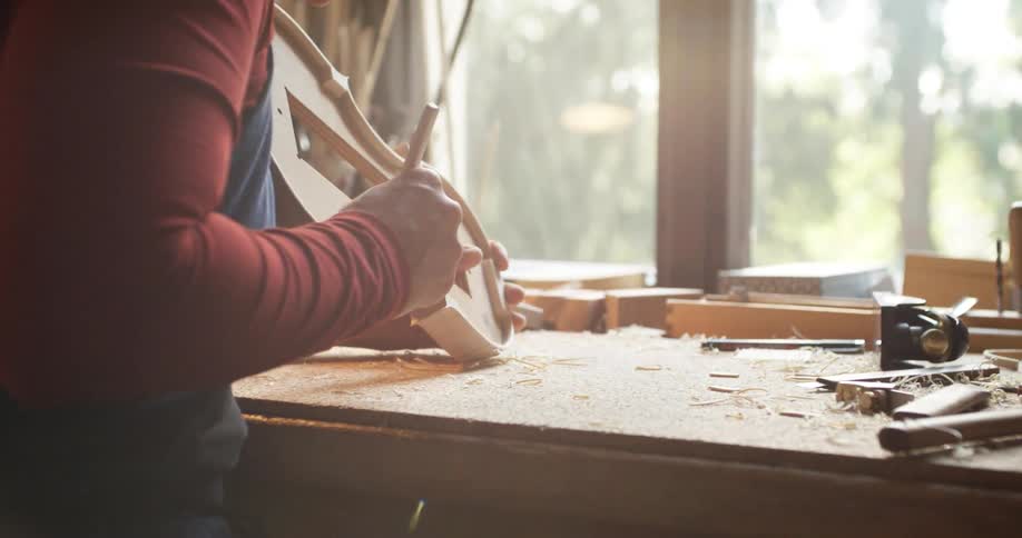 A man is carving the body of a violin during the day