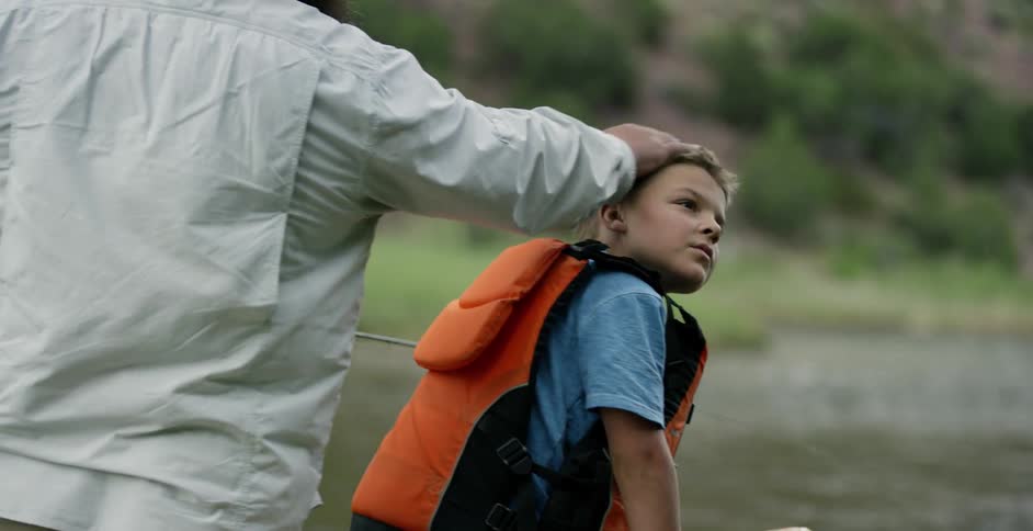 A young boy in a life jacket is standing on a boat and his father is fishing next to him
