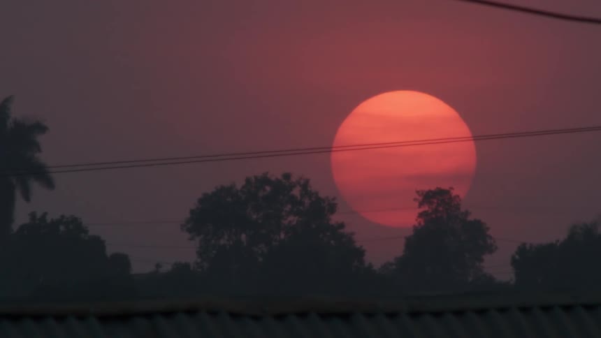 A red sun shines gloomy behind clouds and trees during sunset
