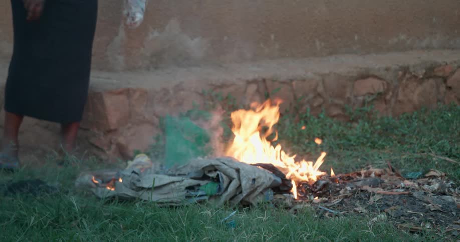 Woman tossing something plastic into a small fire that's burning in a grassy area