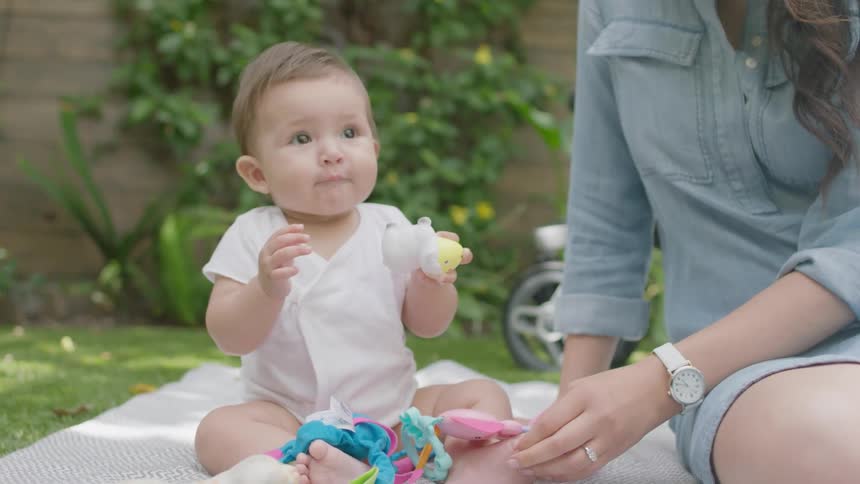 A baby claps as it plays with toys outside with its mother