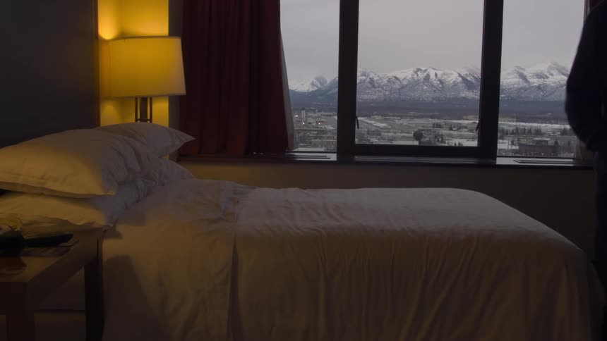 A young man moves around a hotel room with snow capped mountains visible through a window