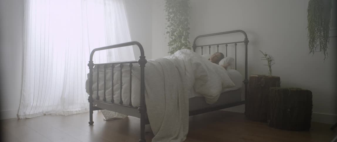 A woman lays in bed as curtains blow in the wind