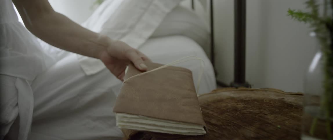 A woman sits on the side of a bed and picks up a book