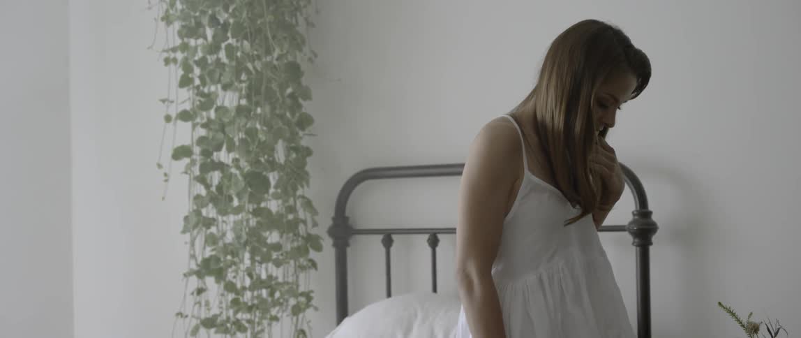 A woman wearing a white dress stands by her bed
