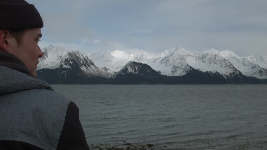 Man standing by the water looking at large mountains with snow on top