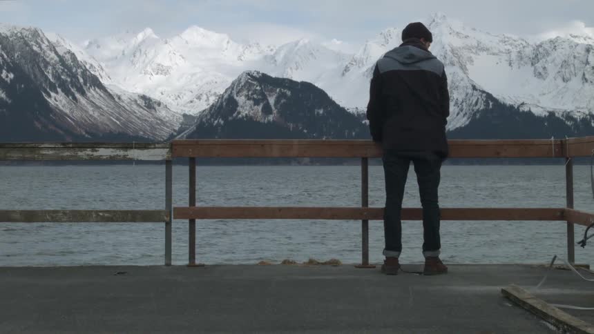 A man stands on a dock looking off towards a range of snowy mountains