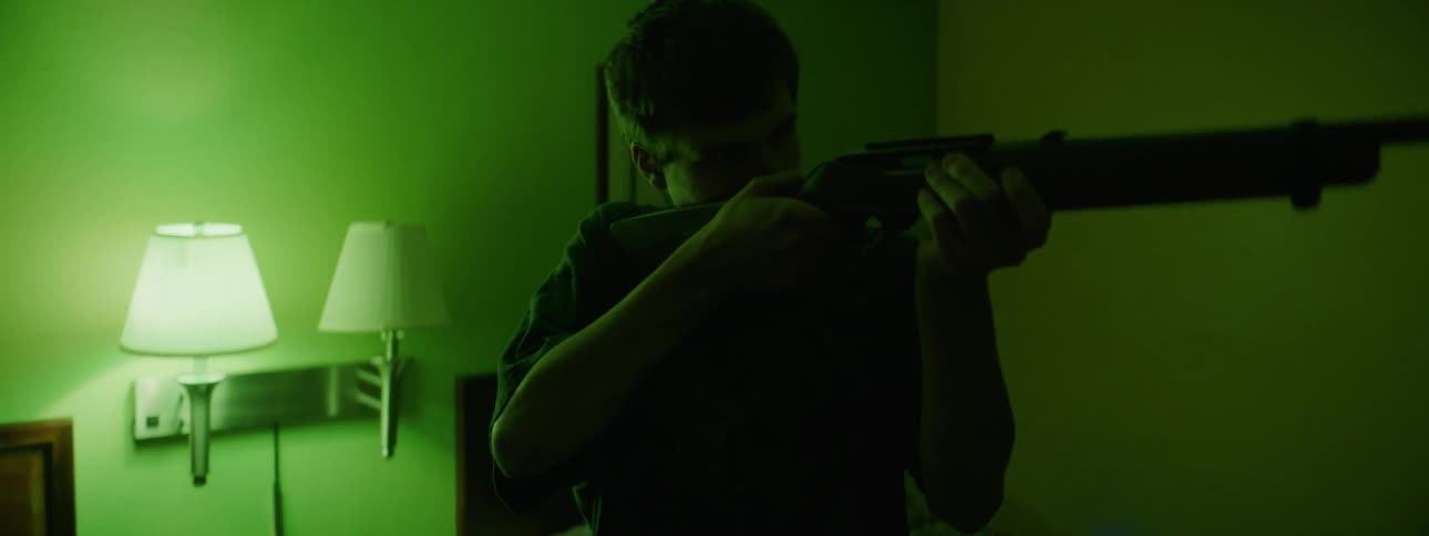 A young man is standing in a hotel room holding a rifle and pointing it at something off screen