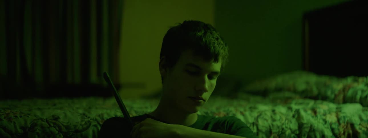 A teenage boy sits next to a bed holding a rifle