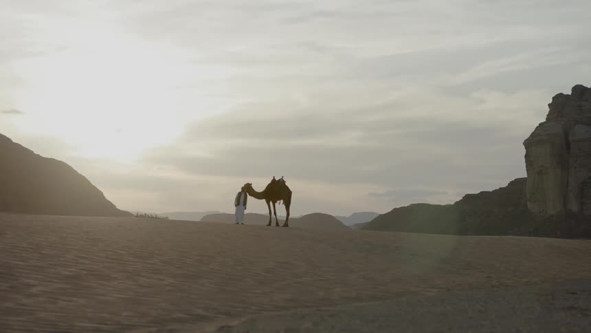 A man is standing next to his camel in the desert,