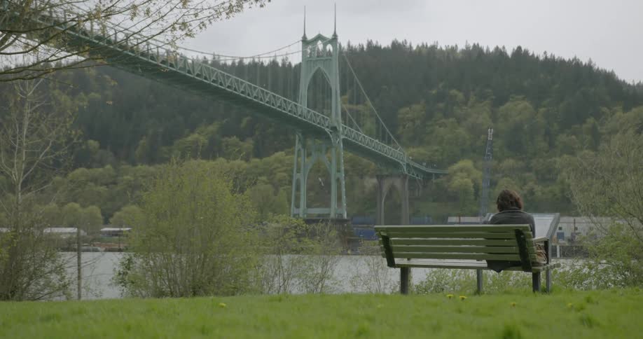 A person sits alone on a bench in a park overlooking a river with a bridge on an overcast day