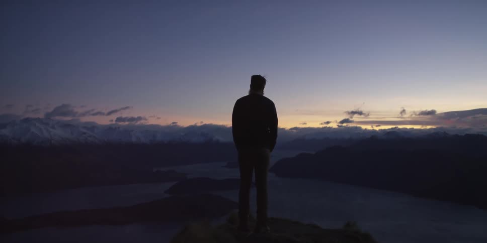 A man stands on a peak overlooking a river at dusk