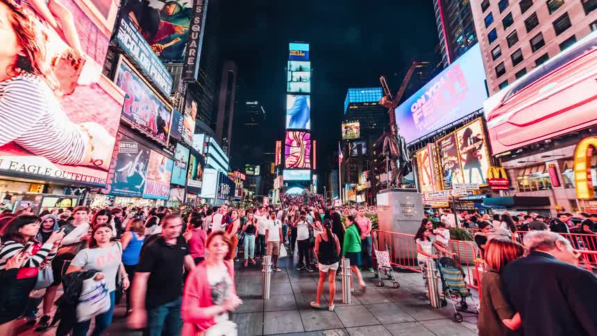 Digital billboards flash as people walk through a city square at night