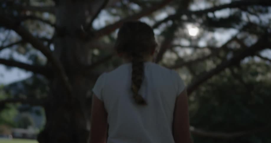 A young girl walks under some big trees in the sunshine