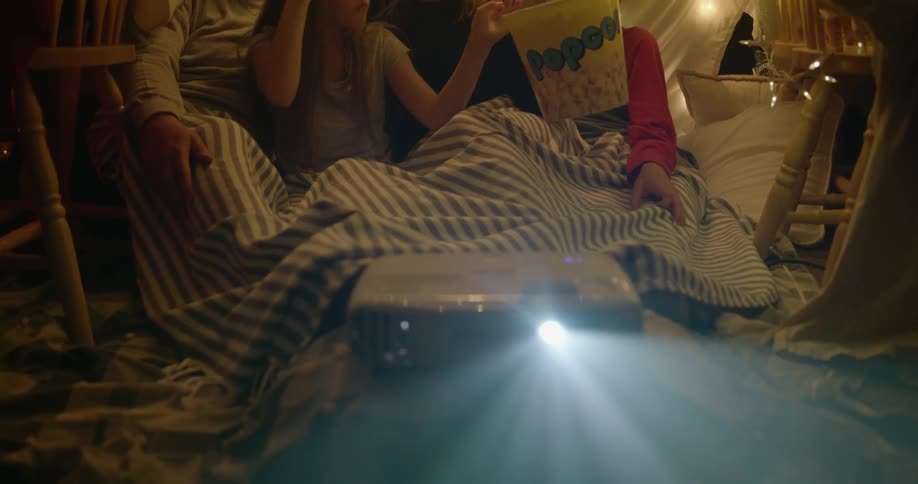 A family is sitting under a blanket eating popcorn and watching movies
