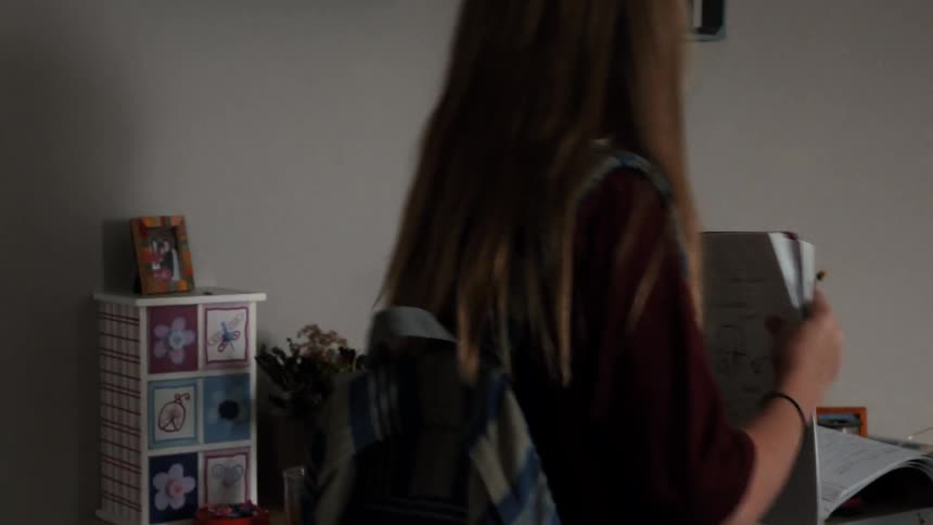 A girl carrying a notebook walks into her bedroom and sits down at her desk