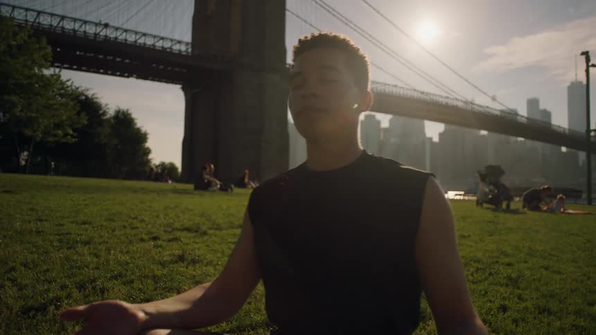 A man is meditating on a field near the bridge