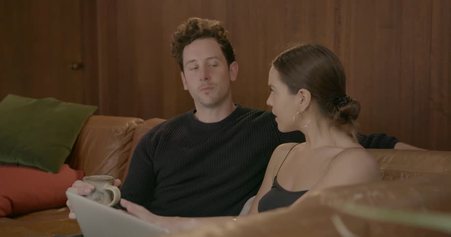 A couple sit on the couch talking and using a laptop