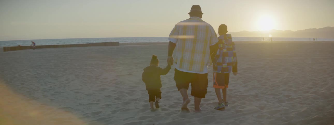 A large man and two small kids are walking on the beach towards the ocean