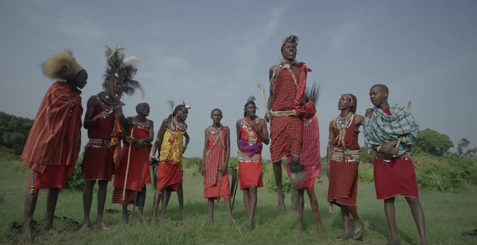 Maasai people wearing costumes stand in a semi circle while one jumps up and down