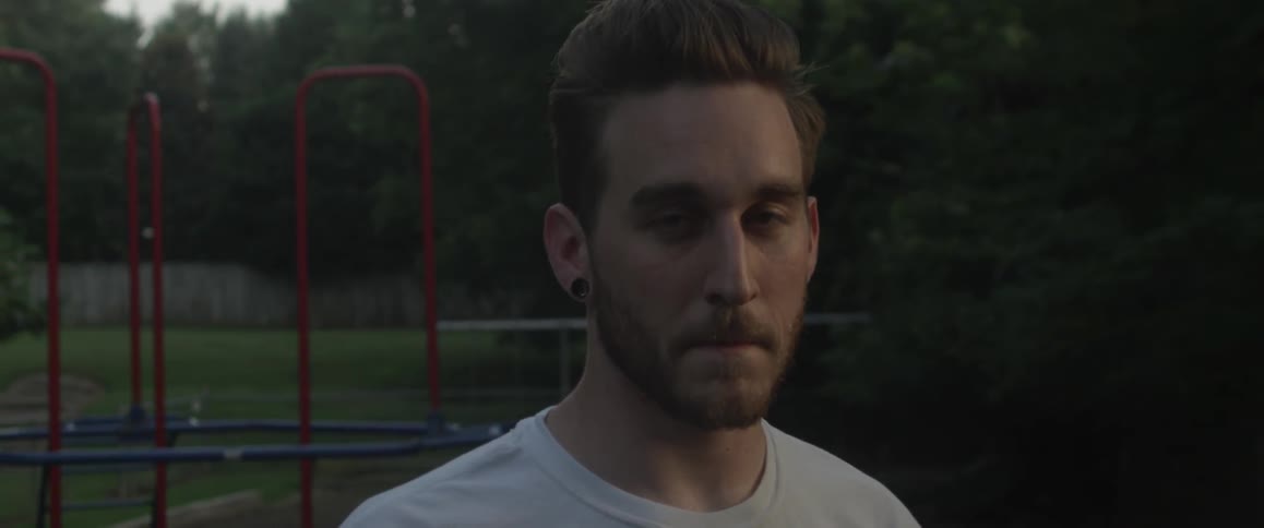 Man wearing a white t shirt is standing outdoors and looking somewhat forward