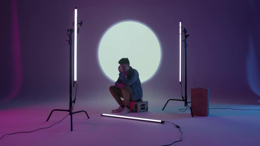 A young man sits on a box in a studio with a light projected behind him