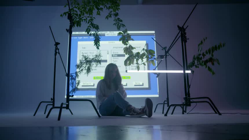 A woman is sitting on a studio floor while computer text displays behind her