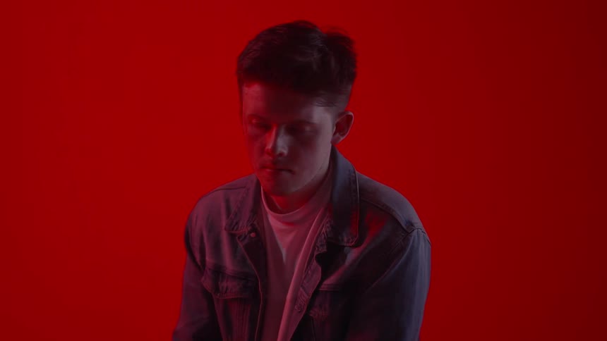 A young man sitting in front of a red wall turns his head and looks down