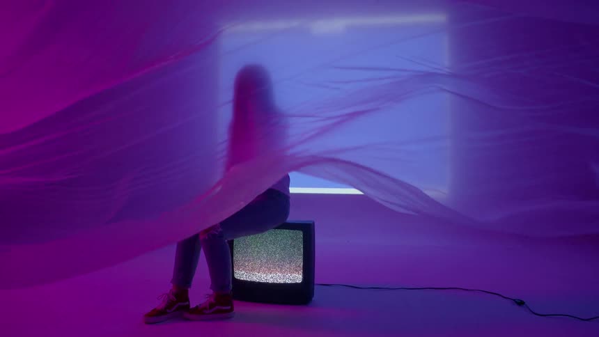 A young woman is sitting on a television as fabric flows in front of her