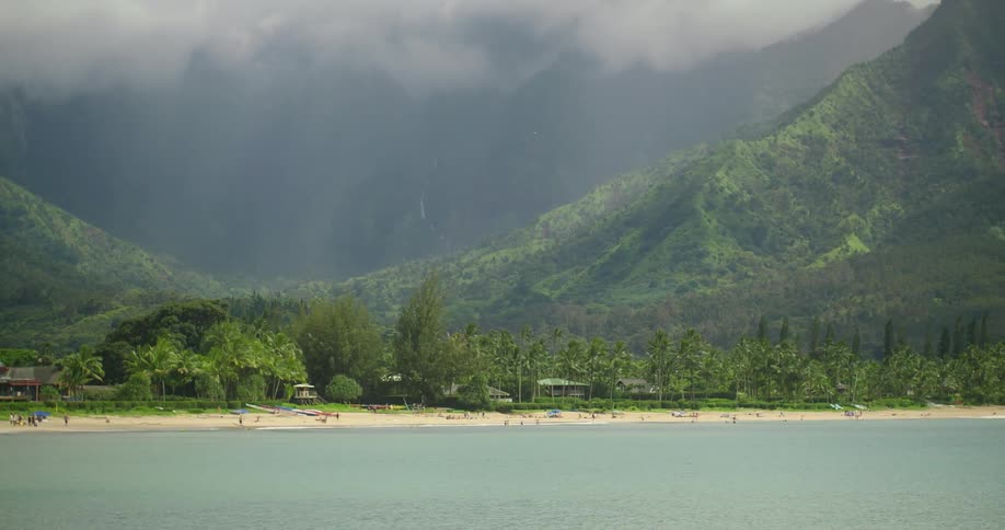 People walk on the beach in front of greenery covered mountains