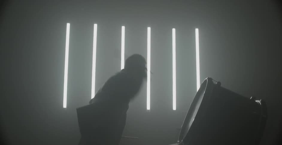 A man is playing drums in a room with a slatted window during the day