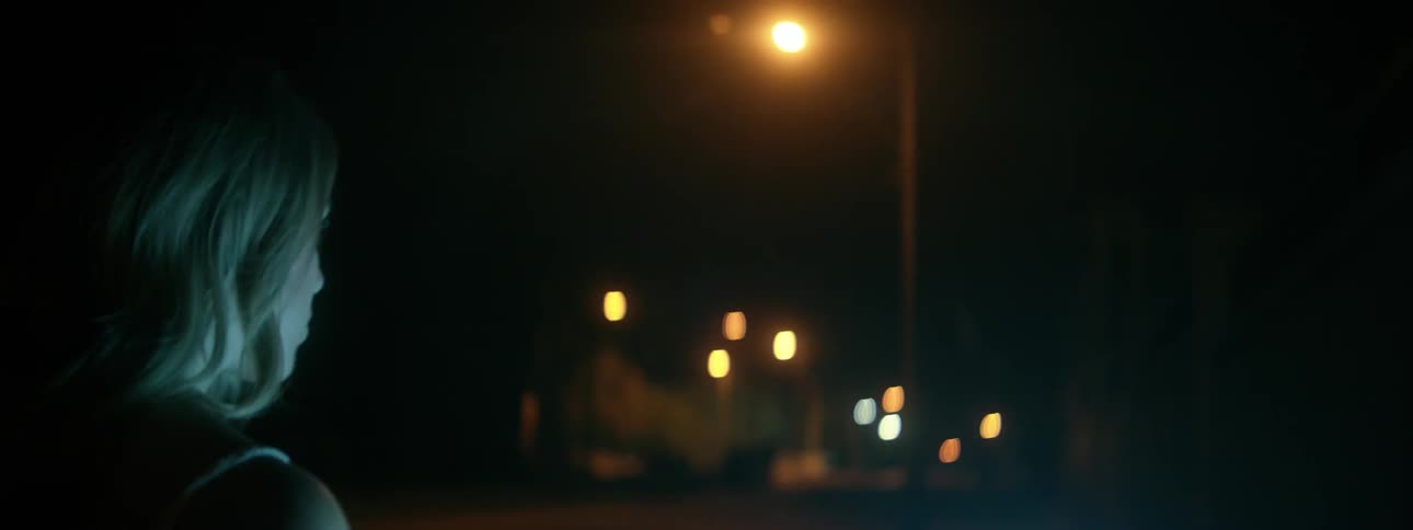 A woman carrying a lightsaber is walking down a street at night