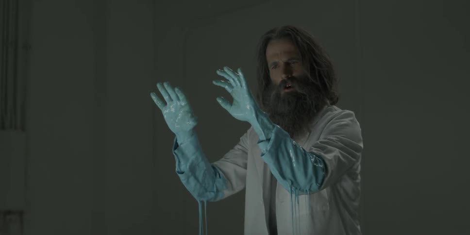 A disheveled man in a lab coat examines his hands that are covered in blue liquid